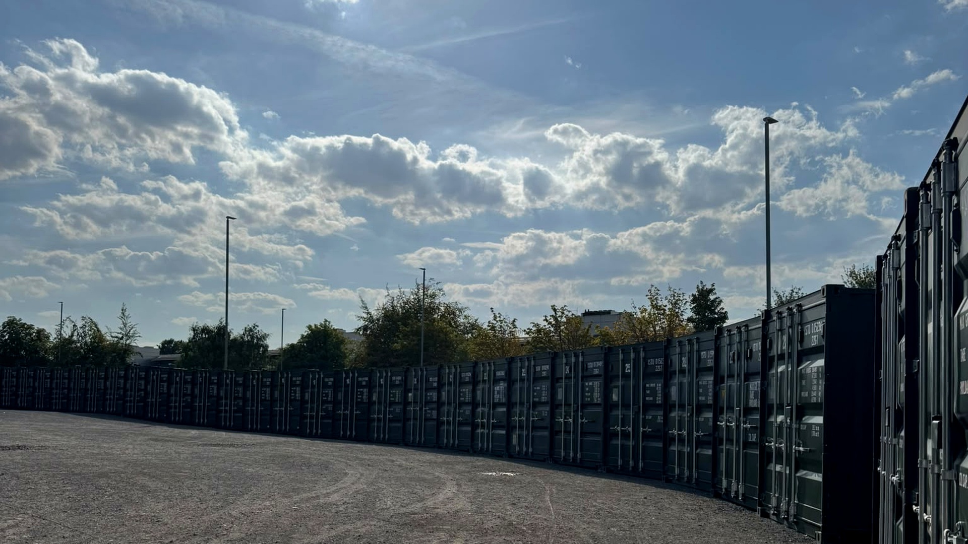 A long curving row of green 20ft storage containers on the gravel yard at MB Storage Leeds in Stourton, under a blue sky